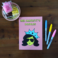 Load image into Gallery viewer, Photo of Her Majesty’s Doodles notebook, featuring a pink cover with the title in yellow and a stylised illustration of an unsmiling woman with long black hair, wearing sunglasses and an extravagant blue crown. Next to the notebook are an assortment of fondant fancies and three felt tip pens.