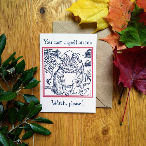 Photograph of a white card on a table next to a brown envelope and leaves of multiple colours. The card features a mediaeval woodcut design of two peasants embracing next to a tree, with a village on a hill in the background. Above in a black Gothic font are the words 