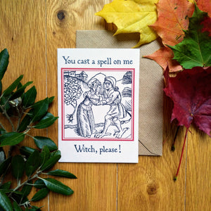 Photograph of a white card on a table next to a brown envelope and leaves of multiple colours. The card features a mediaeval woodcut design of two peasants embracing next to a tree, with a village on a hill in the background. Above in a black Gothic font are the words "You cast a spell on me” and below are the words “Witch, please!”.
