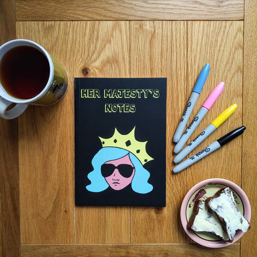 Photo of Her Majesty’s Notes notebook with the title in yellow-outlined letters, featuring a black cover with a stylised illustration of an unsmiling woman with long blue hair, wearing sunglasses and an extravagant yellow crown. Next to the notebook are a mug of black tea, a plate containing buttered toast, and four felt tip pens.