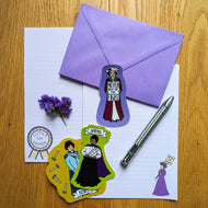 Photo of several stickers, each showing an individual suffragette. One holds a sign saying “Never Give Up The Fight” and another has the caption “Shero” above her head. They are spread out on two blank sheets of paper along with a purple envelope and silver pen.