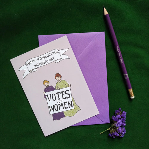 Photo of a greetings card depicting two suffragettes standing together and holding a sign that reads “Votes For Women”. Above is a banner reading 