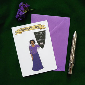 Photo of a greetings card depicting a suffragette in a headscarf and a purple top and skirt, holding a pennant that reads “Votes For Women” with an elephant at the base. Above is a banner reading "Independent Chick" and the background is white. Also pictured besides the card & purple envelope are a silver pen and a sprig of lavender.