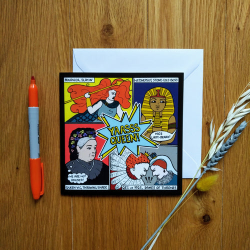 Photo of the card on a white envelope. The image is divided in four, showing a woman with giant red hair and holding a spear (“Boudicca, slayin’”); a pharaoh (“Hatshepsut, stone-cold boss”) scowling as someone out of shot shouts “Nice lady-beard!”; Queen Victoria with a thought bubble that says “We are not amused!” (“Queen Vic, throwing shade”); and Mary, Queen of Scots and Queen Elizabeth I scowling and butting heads (“QE1 vs MQS, Dames of Thrones”). Next to the card is a sprig of wheat and marker pen.