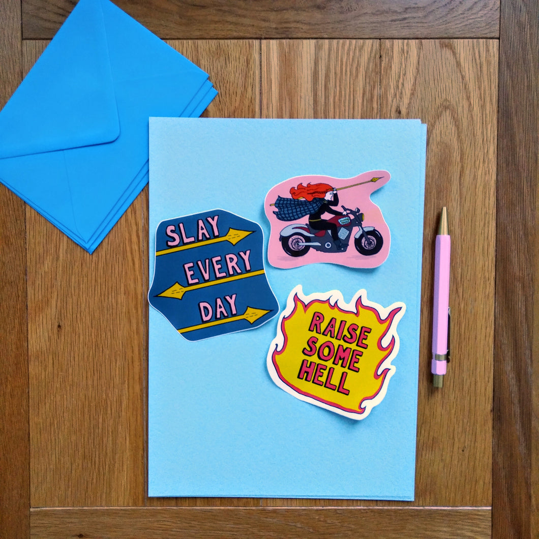 Photo of a blue sheet with three stickers on top. The first has a pink background and shows Boudicca riding a motorbike and wielding a spear overhead. The second says “Slay Every Day” and underlined by spears. The third looks like a fire with “Raise Some Hell” as the text. Next to the stickers is a pink pen and a blue envelope.
