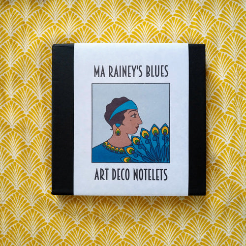 Photo of the notelets bound by a white sleeve, captioned: “Ma Rainey’s Blues Art Deco Notelets.” At the centre is an Art Deco-style illustration of Ma Rainey, a Black woman in a blue dress and headband holding a blue and yellow feather fan.