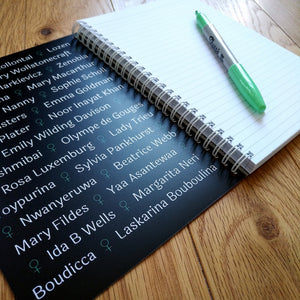 A black spiral-bound notebook, opened, showing a blank page of lined paper and the inside cover, which is black but filed with a list of names of famous women separated by the feminine symbol. A green pen rests on the paper.