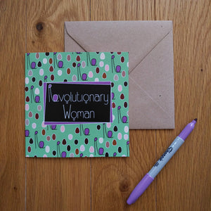 A light turquoise card with minimalist figures of different colours and sizes representing a crowd of women with their fists upraised. The combined head and raised fist resembles the letter “R”, which is used in the caption. In the centre is a black rectangle outlined in purple with the words “Revolutionary Women” in white. The “o”s in the words are turned into feminine symbols. Next to the card is a brown envelope and a purple pen.
