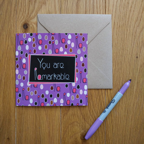 A purple card with minimalist figures of different colours and sizes representing a crowd of women with their fists upraised. The combined head and raised fist resembles the letter “R”, which is used in the caption. In the centre is a black rectangle outlined in purple with the words “You are Remarkable” in white. The “o”s in the words are turned into feminine symbols. Next to the card is a brown envelope and a purple pen.