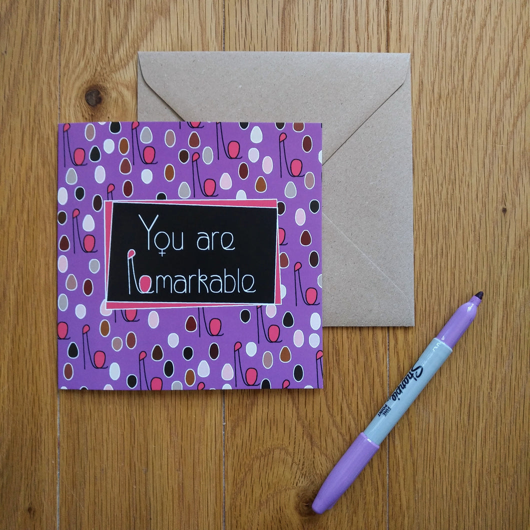 A purple card with minimalist figures of different colours and sizes representing a crowd of women with their fists upraised. The combined head and raised fist resembles the letter “R”, which is used in the caption. In the centre is a black rectangle outlined in purple with the words “You are Remarkable” in white. The “o”s in the words are turned into feminine symbols. Next to the card is a brown envelope and a purple pen.