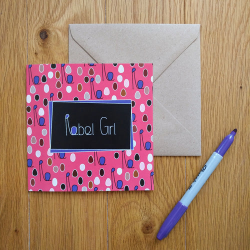 A pink card with covered with minimalist shapes representing a diverse crowd of women with their fists upraised. The combined head and raised fist resembles the letter “R”, which is used in the caption. In the centre is a black rectangle with the words “Rebel Girl” in white. The card is laid on a brown envelope and is next to a marker pen.
