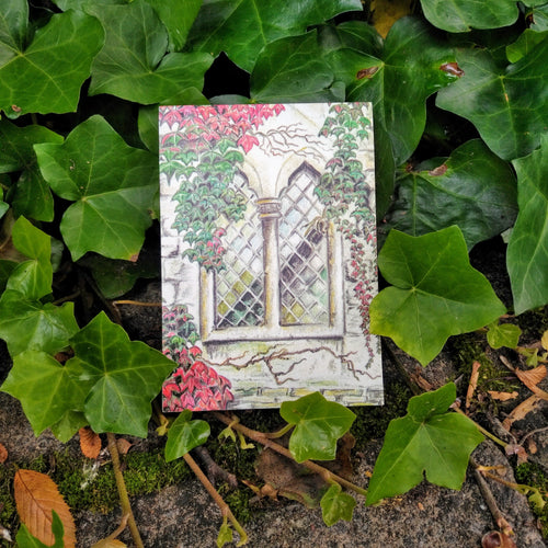 Photo of the notepad, resting on some leaves. The cover shows a gothic window in a stone wall. Around the window are numerous leaves and branches in both green and red. 