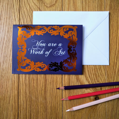 Photo of a greeting card in a dark blue colour with ornate gold leaf styling around the edges. In a white baroque font at the centre is the phrase “You are a Work of Art”. Next to the card is a white envelope and three coloured pencils.