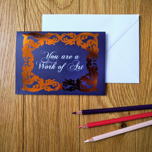 Photo of a greeting card in a dark blue colour with ornate gold leaf styling around the edges. In a white baroque font at the centre is the phrase “You are a Work of Art”. Next to the card is a white envelope and three coloured pencils.