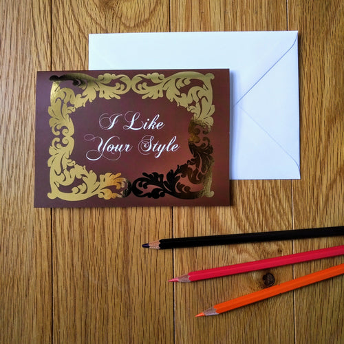 Photo of a greeting card in a burgundy colour with ornate gold leaf styling around the edges. In a white baroque font at the centre is the phrase “I Like Your Style”. Next to the card is a white envelope and three coloured pencils.