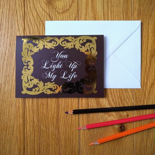 Photo of a greeting card in a dark red-brown colour with ornate gold leaf styling around the edges. In a white baroque font at the centre is the phrase “You Light Up My Life”. Next to the card is a white envelope and three coloured pencils.