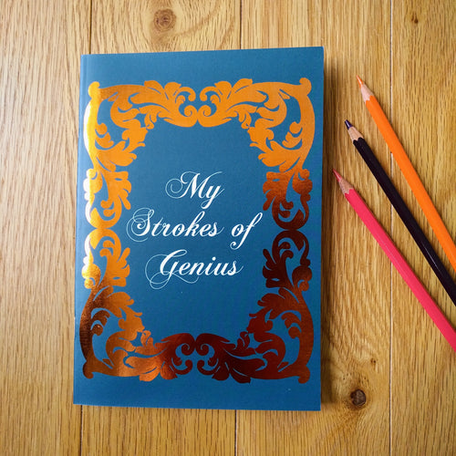 Photo of the notebook, which is blue with a Baroque gold-foil design around the central caption which reads: “My Strokes of Genius”. Next to the notebook are three coloured pencils.