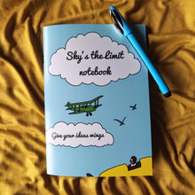 Load image into Gallery viewer, Photo of a light blue notebook. The cover shows an illustrated green biwing plane, and in the foreground we see a yellow plane’s cockpit with an aviator inside. The text in two separate clouds says “Sky’s the limit notebook” and “Give your ideas wings”. Next to the notebook is a blue pen.