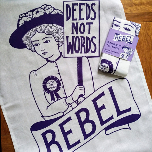 Photo of a white tea towel with an illustration and text, all in purple. The image is of a suffragette wearing a hat with flowers, a rosette, and holding a sign that says “Deeds Not Words”. Beneath her is a banner that says “Rebel”. A folded version of the tea towel with a purple cardboard slip is also shown with the same artwork, and the text “Suffragette Tea Towel” and “Because Housework Is Political”.