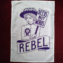 Load image into Gallery viewer, Photo of a white tea towel with an illustration and text, all in purple. The image is of a suffragette wearing a hat with flowers, a rosette, and holding a sign that says “Deeds Not Words”. Beneath her is a banner that says “Rebel”.