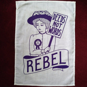 Photo of a white tea towel with an illustration and text, all in purple. The image is of a suffragette wearing a hat with flowers, a rosette, and holding a sign that says “Deeds Not Words”. Beneath her is a banner that says “Rebel”.