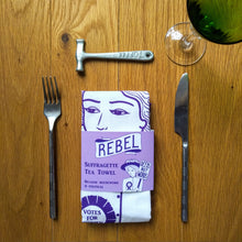 Load image into Gallery viewer, Photo a folded white tea towel with an illustration and text, all in purple. The image is of a suffragette wearing a hat with flowers, a rosette, and holding a sign that says “Deeds Not Words”. Beneath her is a banner that says “Rebel”. The tea towel is in a purple cardboard slip is also shown with the same artwork, and the text “Suffragette Tea Towel” and “Because Housework Is Political”. A knife and fork are on either side, and a toffee hammer is above the tea towel. A wine glass is just in shot.