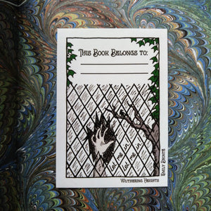 Photo of the bookplate pasted into a book. The bookplate shows an illustrated ghostly hand smashing through a window next to a gnarled dead tree. On the panes are written: “Let me in! Let me in!”. The borders of the plate say “Wuthering Heights” and “Emily Brontë” and there is a space at the top that says “This Book Belongs To” with two dotted lines to fill in the name, all framed by ivy.