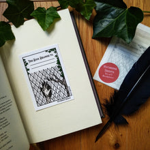 Load image into Gallery viewer, Photo of the bookplate pasted into a book. The bookplate shows an illustrated ghostly hand smashing through a window next to a gnarled dead tree. On the panes are written: “Let me in! Let me in!”. The borders of the plate say “Wuthering Heights” and “Emily Brontë” and there is a space at the top that says “This Book Belongs To” with two dotted lines to fill in the name, all framed by ivy. Next to the book is a quill and a sealed pack of the book plates.