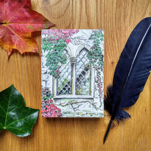 Load image into Gallery viewer, Photo of the notepad, resting on a desk next to a quill and two autumn leaves. The cover shows a gothic window in a stone wall. Around the window are numerous leaves and branches in both green and red.
