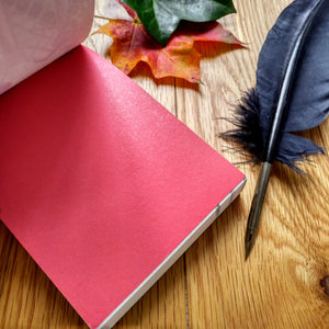Photo of the open notepad, resting on a desk next to two autumn leaves and a black quill pen. The inside leaf of the notebook is entirely red.