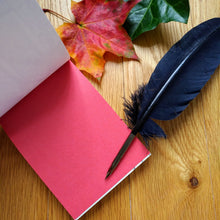 Load image into Gallery viewer, Photo of the open notepad, resting on a desk next to two autumn leaves. The inside leaf of the notebook is entirely red. The nib of the quill rests on the page.