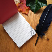 Load image into Gallery viewer, Photo of the open notepad, resting on a desk next to two autumn leaves and a black quill pen. The notepad is open on a blank lined page, in the bottom corner of which is an illustration of some ivy.