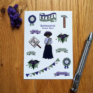 Photo of a sheet showing 14 stickers, all suffragette-themed, including a suffragette, slogans, signs, a hammer, and more. Next to the sheet are some purple flowers and a silver pen.
