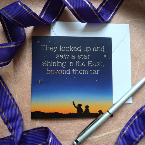 Photo of a card that shows a dark night’s sky over the sunset. We see the silhouette of two figures, one pointing to the sky. Written in the sky in gold foil are the words “They looked up and saw a star Shining in the East, beyond them far”. Next to the card is a white envelope, silver pen, and a curling purple ribbon.