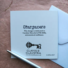 Load image into Gallery viewer, Photo of the back of the card. Shows the Clavis &amp; Claustra key logo and text that says “Stargazers” and “This design is dedicated to Caroline Herschel (1750-1848), astronomical trailblazer.”