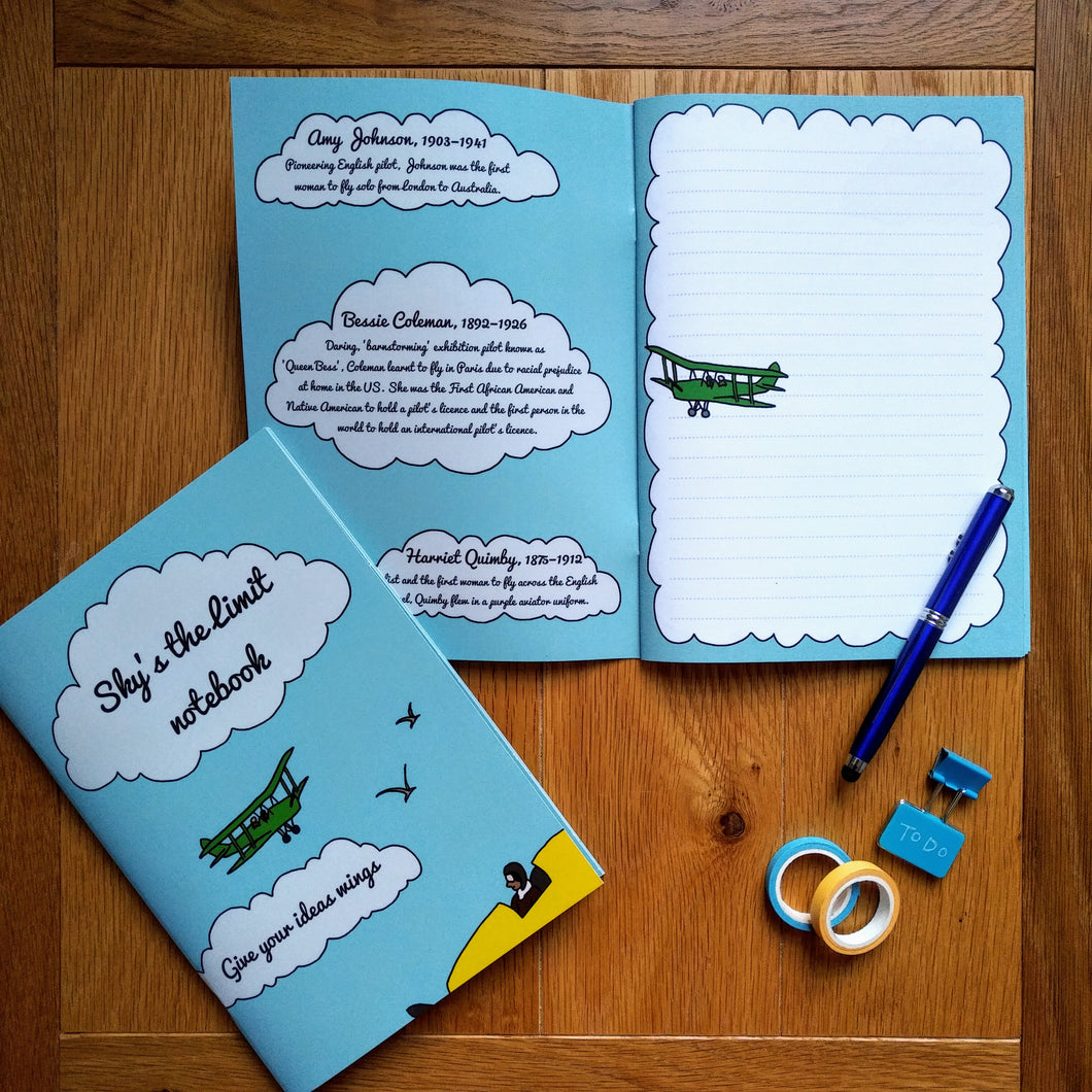 Photo of the notebook both closed and open. The notebook is light blue with an illustrated green biwing plane, and in the foreground we see a yellow plane’s cockpit with an aviator inside. The text in two separate clouds says “Sky’s the limit notebook” and “Give your ideas wings”. The inside has a blank lined page with an illustrated plane, and the inside cover shows numerous female aviator biographies inside clouds. Next to the notebook is a pen, some gift tape, and some document clips.