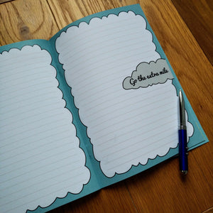 Photo of the open notebook, showing blank lined pages with light blue fringes to make the pages resemble clouds and “Go the extra mile” written in a gray cloud. A pen is resting on the page.
