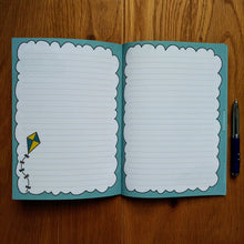 Load image into Gallery viewer, Photo of the open notebook, showing blank lined pages with light blue fringes to make the pages resemble clouds. A blue and yellow diamond-shaped kite rises from the bottom left of the left page. A pen is resting next to the notebook.
