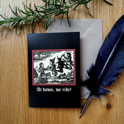 A black coloured card featuring a mediaeval woodcut artwork depicting witches and demons on broomsticks. Beneath it the caption reads 'At dawn, we ride!'. Pictured with a brown envelope, black feather and sprig of rosemary.