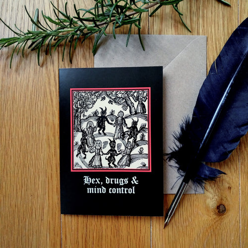 Photo of a greetings card featuring a black-and-white mediaeval woodcut artwork on a black background, depicting witches and demons joining hands and dancing in a circle. Beneath it the caption reads 'Hex, drugs & mind control' in a white Gothic typeface. Next to the card is a brown envelope, quill, and plant branch.