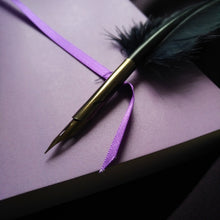 Load image into Gallery viewer, Photo of the inside of the notebook showing a page with a purple ribbon draped across it. The nib of a quill-styled pen rests on top of it.