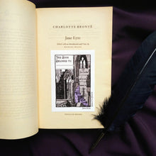 Load image into Gallery viewer, Photo of the Jane Eyre bookplate, which illustrates gothic windows and stairs and “I am no bird and no net ensnares me” carved into the wall. A raven sits on a broken pillar. In the top left is a space to write starting with “This Book Belongs To…”. The bookplate is pictured resting on the title page of Jane Eyre next to a black quill.