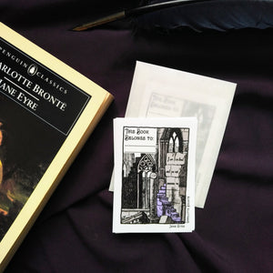 Photo of the Jane Eyre bookplate, which illustrates gothic windows and stairs and “I am no bird and no net ensnares me” carved into the wall. A raven sits on a broken pillar. In the top left is a space to write starting with “This Book Belongs To…”. The bookplate is pictured next to the Penguin Classics edition of Jane Eyre.