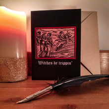 Load image into Gallery viewer, Photograph of a black card standing on a table next to a candle, brown envelope and quill. The card features a mediaeval woodcut design depicting a group of people talking and one woman riding off on a winged demon. Below in a white Gothic font are the words &quot;Witches be trippin&#39;&quot;.