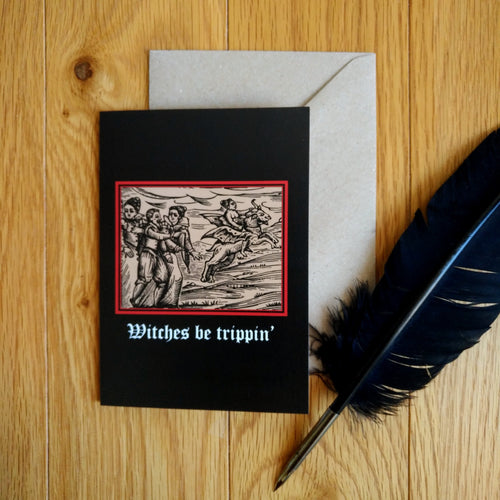 Photograph of a black card on a table next to a brown envelope and quill. The card features a mediaeval woodcut design depicting a group of people talking and one woman riding off on a winged demon. Below in a white Gothic font are the words 
