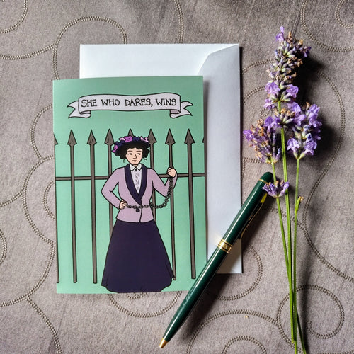 Photo of a pale green greeting card showing a suffragette chained to a spiked iron fence. The caption reads “She who dares, wins”. Next to the card is a green fountain pen and a sprig of lavender.