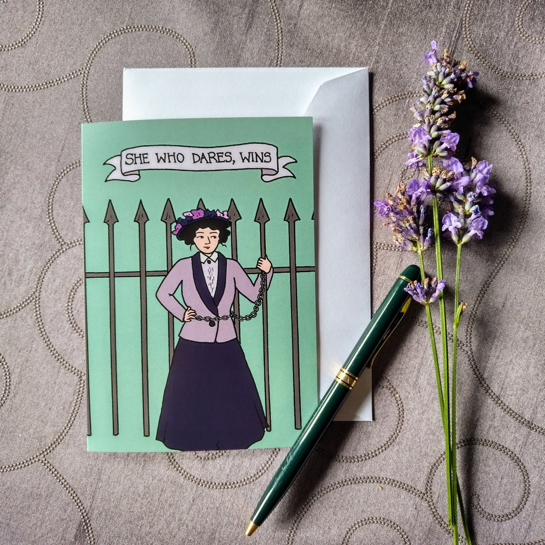 Photo of a pale green greeting card showing a suffragette chained to a spiked iron fence. The caption reads “She who dares, wins”. Next to the card is a green fountain pen and a sprig of lavender.
