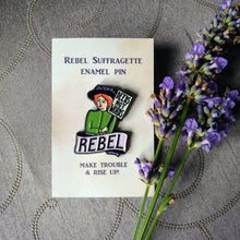 Load image into Gallery viewer, Photo of an enamel pin, depicting a suffragette with ginger hair, a black hat, and a green top. She holds a sign that says “Deeds Not Words” and has a banner beneath her that says “Rebel” in black text on a pink background. The backing card says “Rebel Suffragette Enamel Pin” and “Make Trouble &amp; Rise Up!”. Next to the pin is a sprig of lavender.