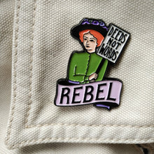 Load image into Gallery viewer, Photo of an enamel pin, depicting a suffragette with ginger hair, a black hat, and a green top. She holds a sign that says “Deeds Not Words” and has a banner beneath her that says “Rebel” in black text on a pink background.