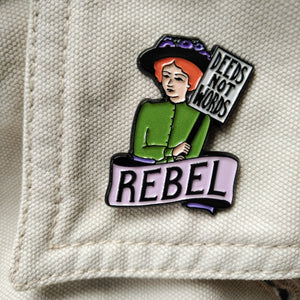 Photo of an enamel pin, depicting a suffragette with ginger hair, a black hat, and a green top. She holds a sign that says “Deeds Not Words” and has a banner beneath her that says “Rebel” in black text on a pink background.